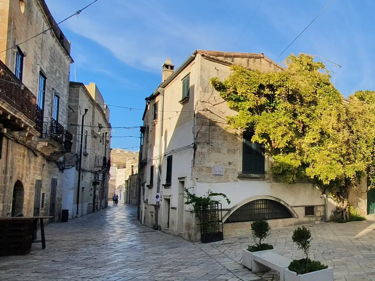 Largo San Marco a Francavilla Fontana: case in pietra calcarea attorno a una piazzetta lastricata, un grande albero e una strada che sale in fondo.